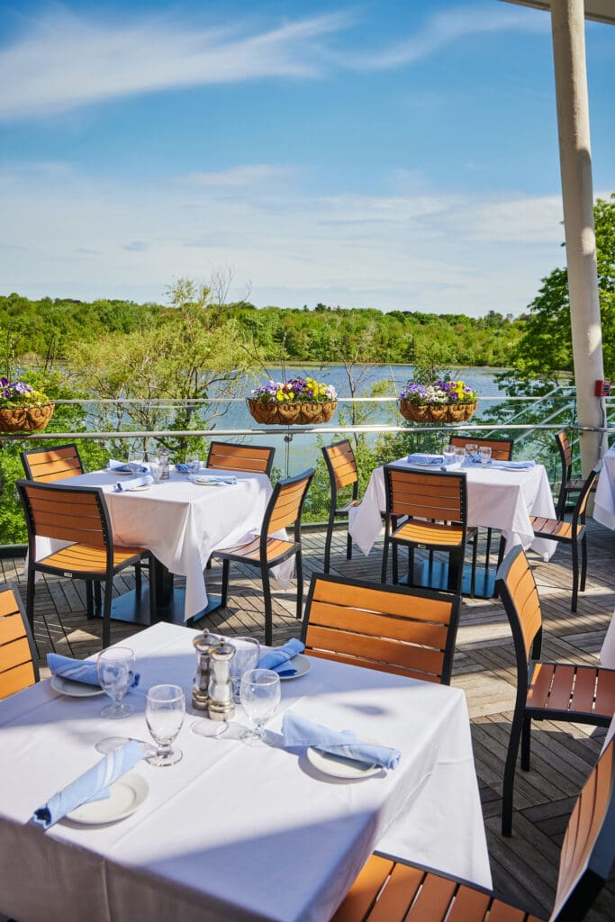 An outdoor restaurant with wooden chairs and white tablecloth-covered tables. Tables are set with glassware, plates, and napkins. Flower pots with colorful blooms hang along the edge of the seating area, with a scenic view of a lake and greenery in the background.