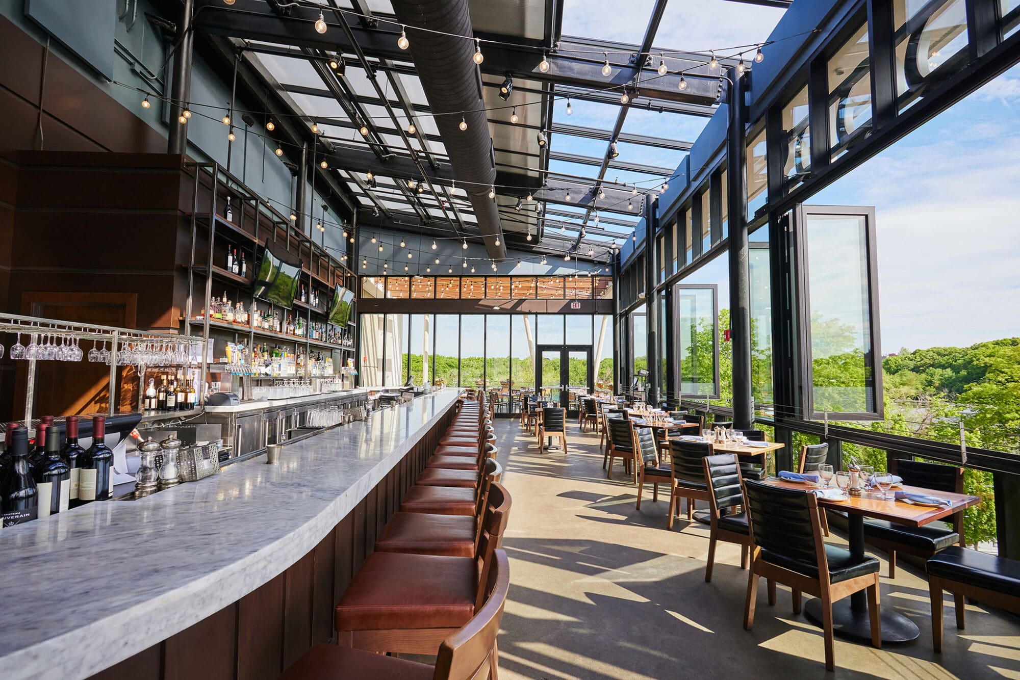 A bright, modern restaurant interior with an expansive glass roof and windows, offering scenic views of lush greenery. The space features a long, white marble bar with leather bar stools, wooden dining tables, and chairs, all set up in a spacious, open layout.