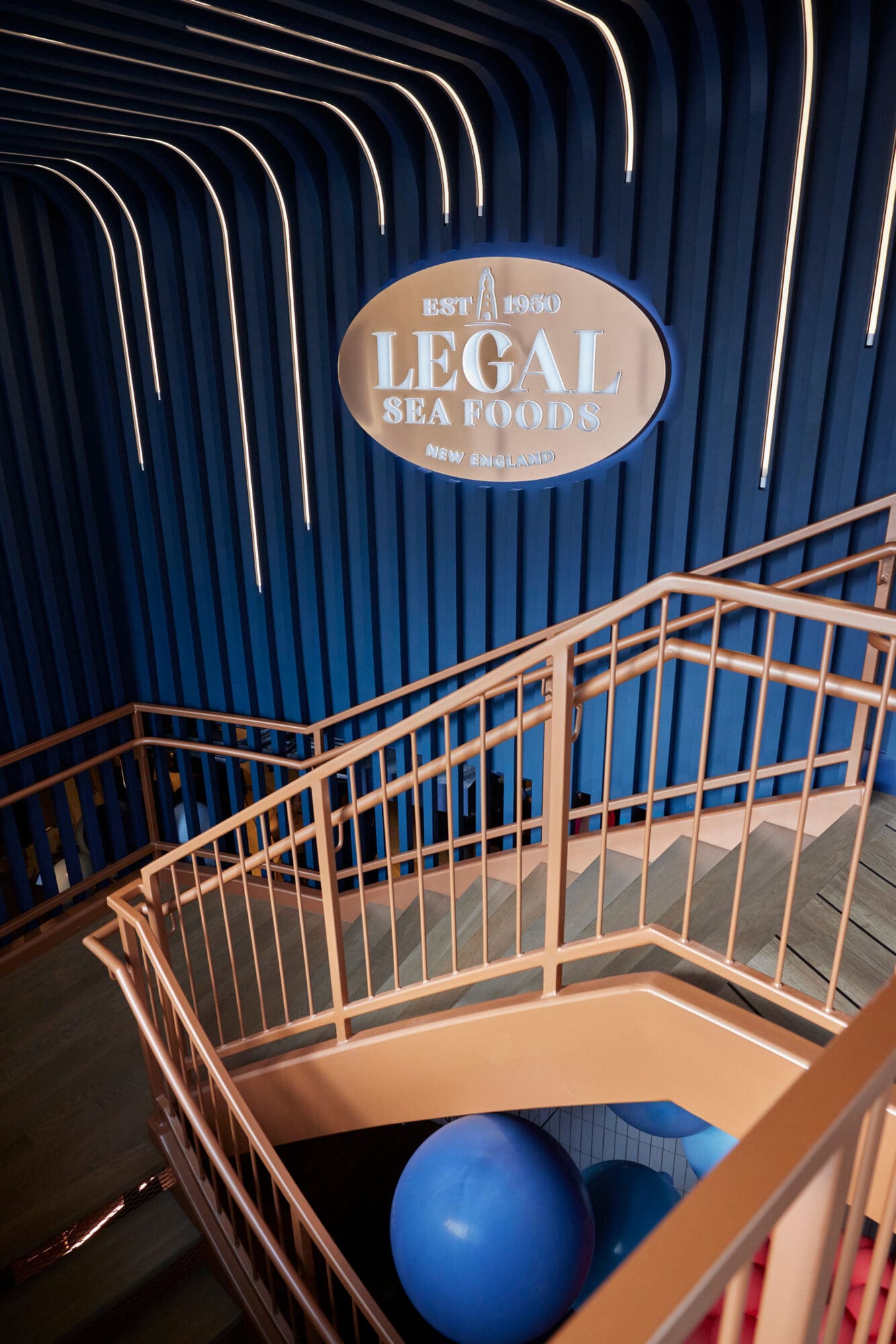 A wooden staircase with metal railings leads downwards in a dimly lit interior. On the blue wall above, a large sign reads 