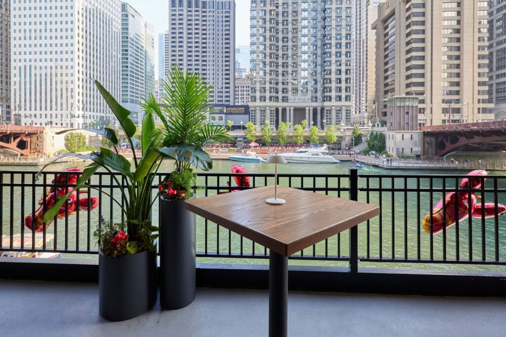 A wooden table with a single coaster is on a patio beside a black railing, overlooking a river. Two black planters with tall green plants and red flowers flank the table. High-rise buildings and a river with boats are visible in the background under a clear sky.