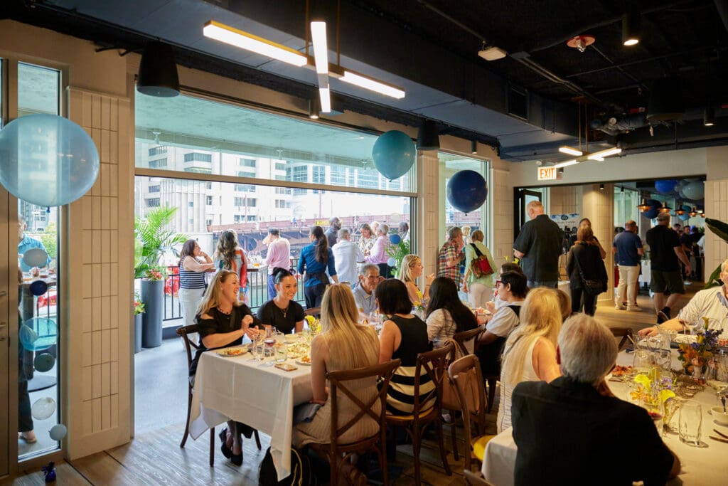 A group of people at a social gathering are seated at a long table indoors, enjoying a meal with drinks and decorations, while others in the background stand and socialize on a patio with a city skyline view. Blue balloons are visible, adding to the festive atmosphere.