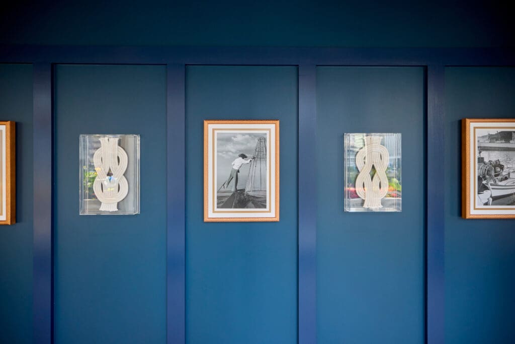 A deep blue wall adorned with three framed artworks. The center frame features a black-and-white photograph of a person on a boat, while the other two frames contain abstract sculptures with vertical, ribbon-like shapes.