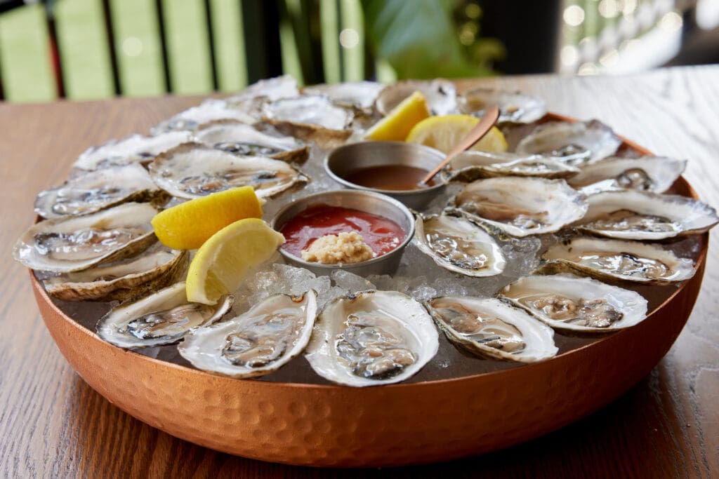 A large platter of oysters on ice is served with lemon wedges, cocktail sauce, and a small container of horseradish. The oysters are arranged in a circular pattern on a copper tray, ready to be enjoyed.