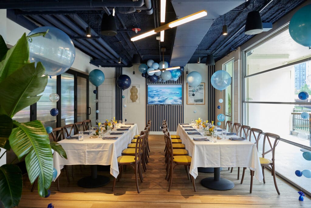 A dining setup in a modern room with long tables covered in white tablecloths, wooden chairs with yellow cushions, and blue-themed decorations. Balloons, including white and various shades of blue, are arranged around the room, adding a festive atmosphere.