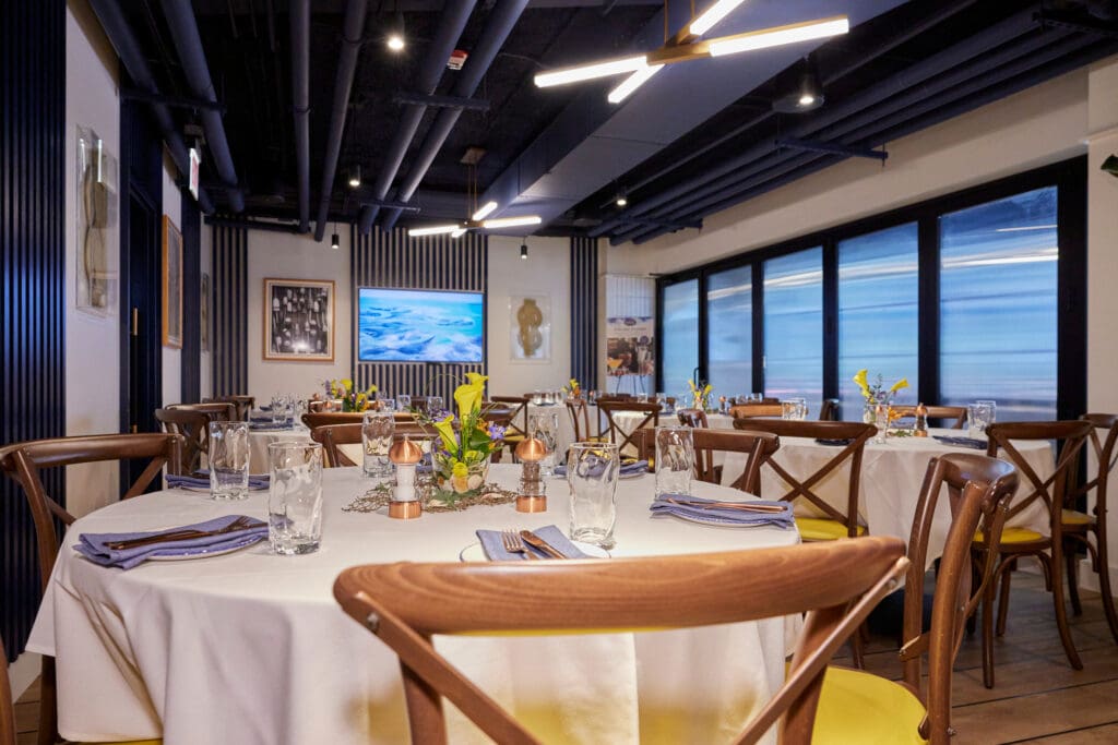 A well-lit, modern restaurant dining area with several round tables covered in white tablecloths. Each table is set with glassware, napkins, and small flower arrangements. The wooden chairs have cushioned seats, and the walls are adorned with artwork and large windows.