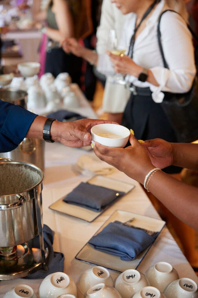 Two people exchange a bowl of soup at a buffet table. In the background are other attendees, some holding plates and cups. The table is set with napkins, upside-down cups labeled with names, and a large soup pot. The scene is lively with social interaction.