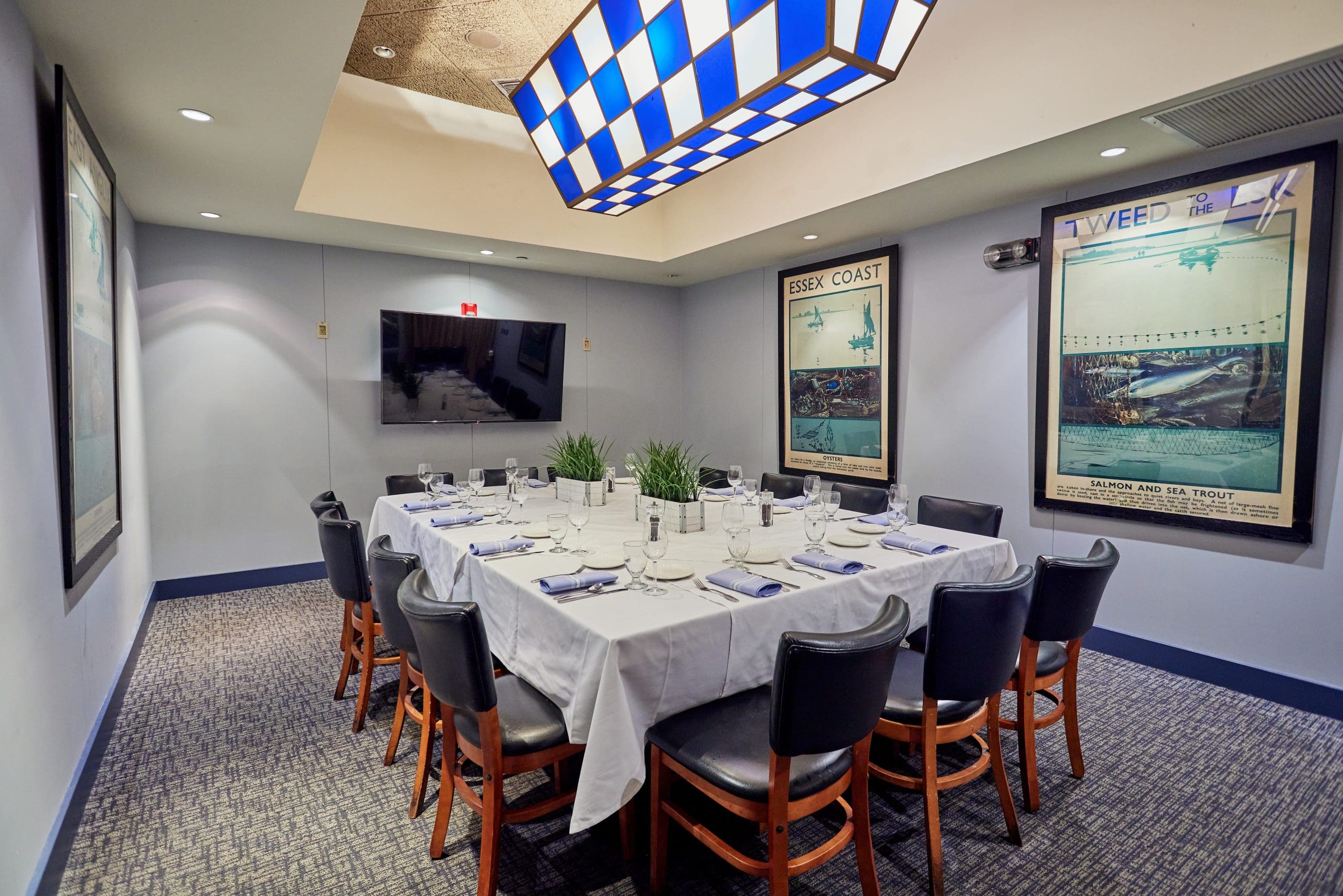 A small, elegantly decorated conference room with a rectangular table covered with a white tablecloth and set with eight black chairs. The walls are adorned with vintage posters, and a large flat-screen TV is mounted on the far wall. A blue and white checkered light fixture hangs from the ceiling, and potted plants decorate the table.