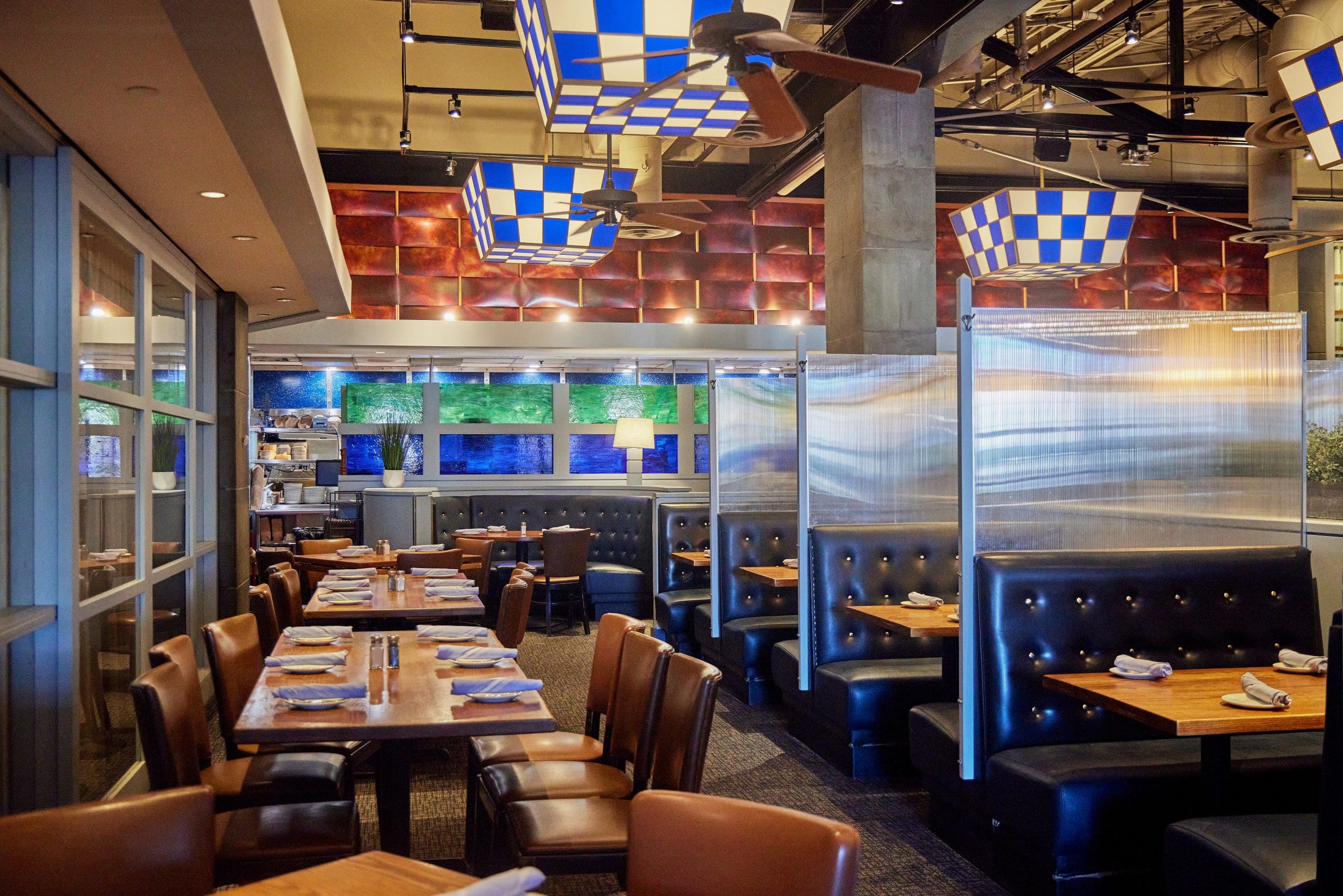 A modern restaurant interior with wooden tables, brown leather chairs, and cushioned black booths. Tables are set with white napkins and cutlery. The lighting is from geometric pendant fixtures, and the decor features metal dividers and a colorful ceiling design.