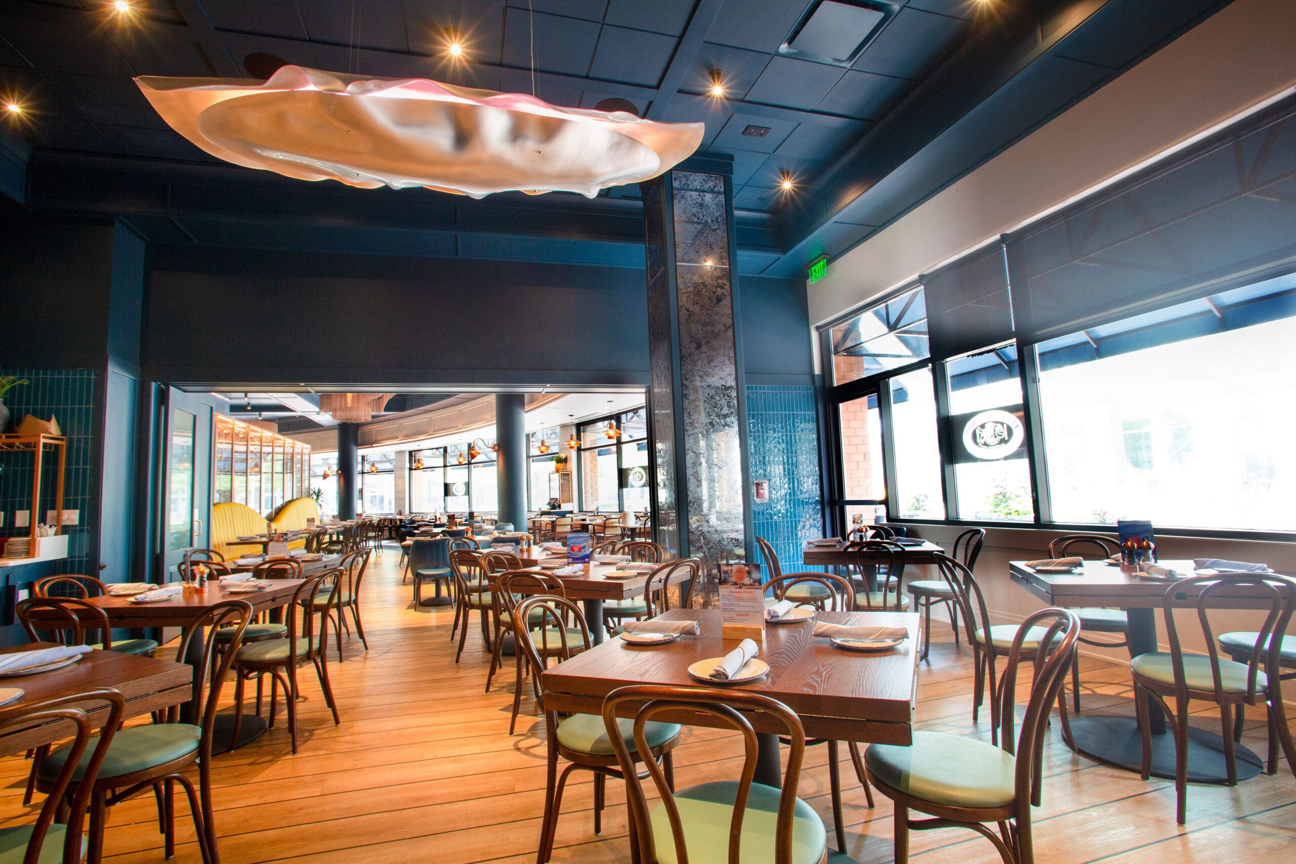 A modern restaurant with wooden floors and furniture. The tables are neatly set with plates and rolled napkins. There is a large decorative ceiling feature resembling a curved shell. Natural light streams in through large windows on the right side.