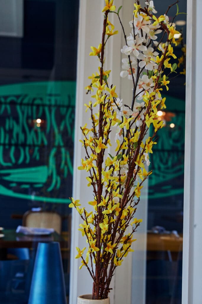 A tall vase with branches of yellow and white flowers is placed indoors. The background features a window with reflections of a green mural and a hint of a dining area with chairs and tables. The setting appears to be a restaurant or cafe.