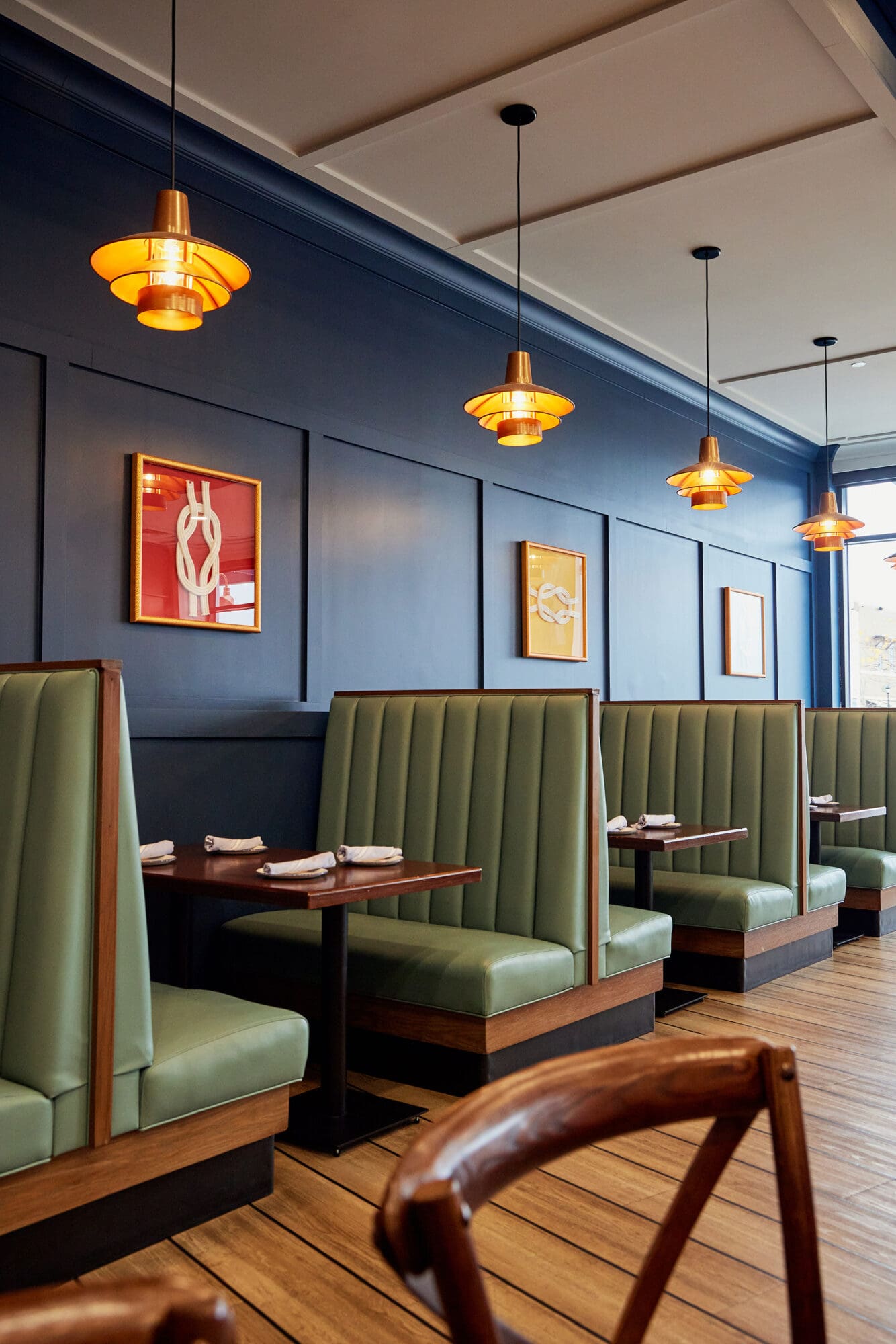 A cozy, modern restaurant interior featuring teal leather booth seating and dark wooden tables. Warm overhead pendant lights hang along the navy blue paneled wall adorned with minimalistic framed art. The floor is lined with wooden planks, and napkins are neatly arranged on the tables.