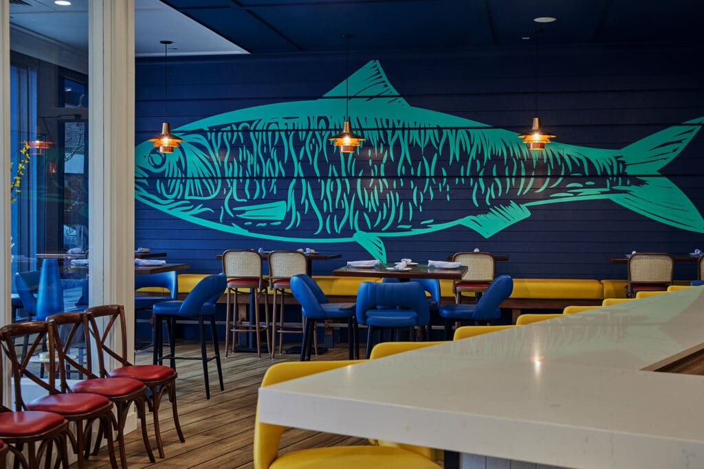 A modern restaurant interior featuring a large blue-green fish mural on a navy blue wall. The seating includes red and yellow cushioned stools, blue chairs, and wooden chairs. Three pendant lights hang above the dining area, enhancing the relaxed atmosphere.