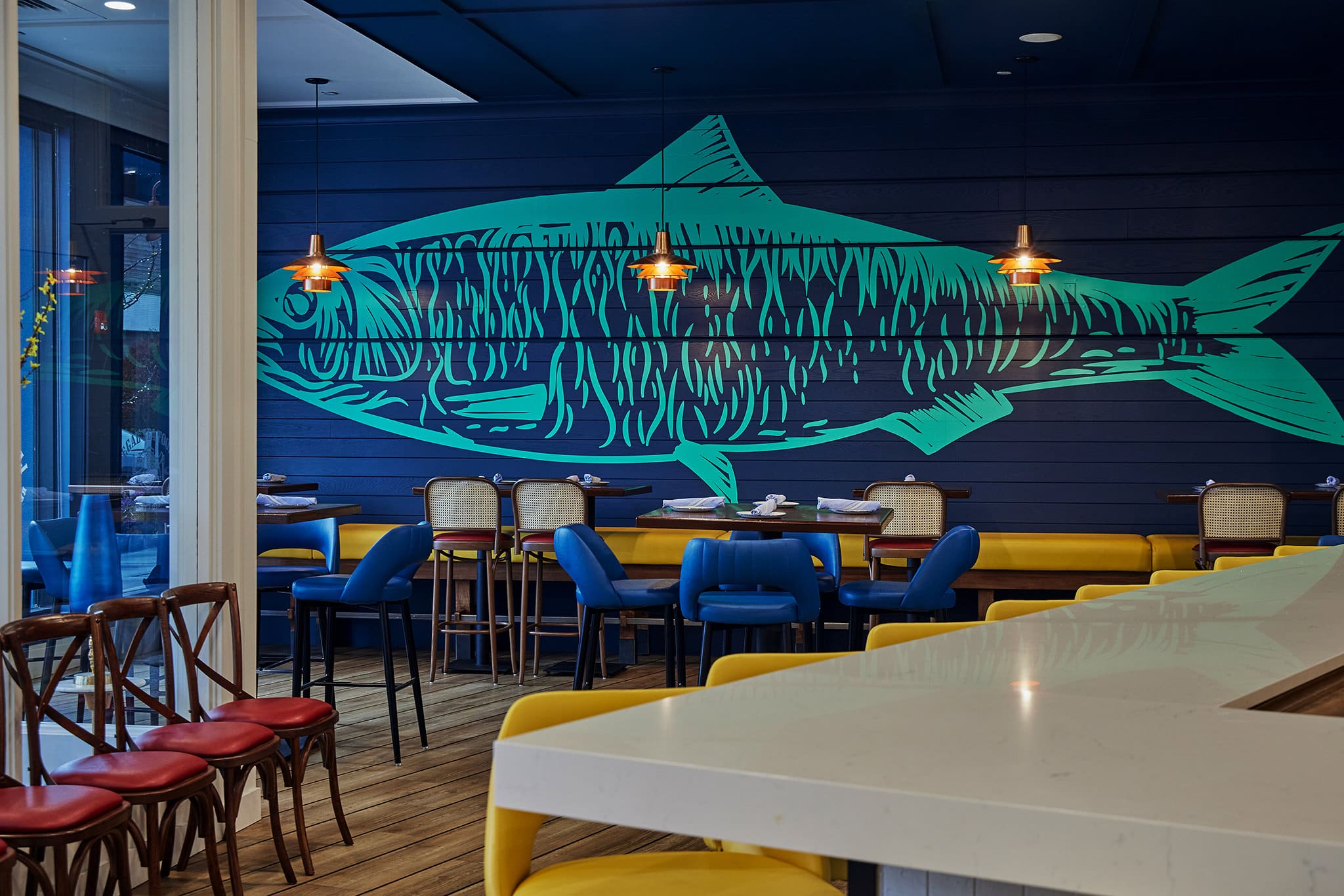 A modern restaurant interior featuring a large blue-green fish mural on a navy blue wall. The seating includes red and yellow cushioned stools, blue chairs, and wooden chairs. Three pendant lights hang above the dining area, enhancing the relaxed atmosphere.