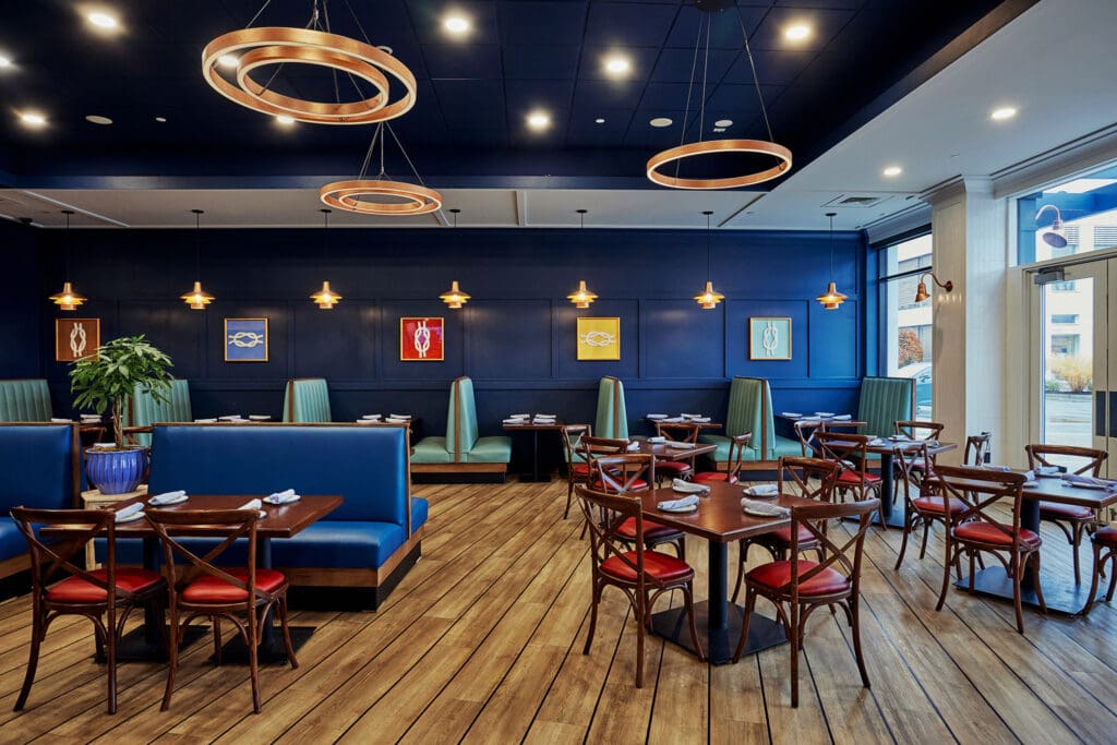 A modern, well-lit restaurant interior with blue walls, wooden flooring, and a mix of booth seating and tables with chairs. Art pieces are hung on the walls, and stylish light fixtures hang from the ceiling. Tables are set with white napkins.