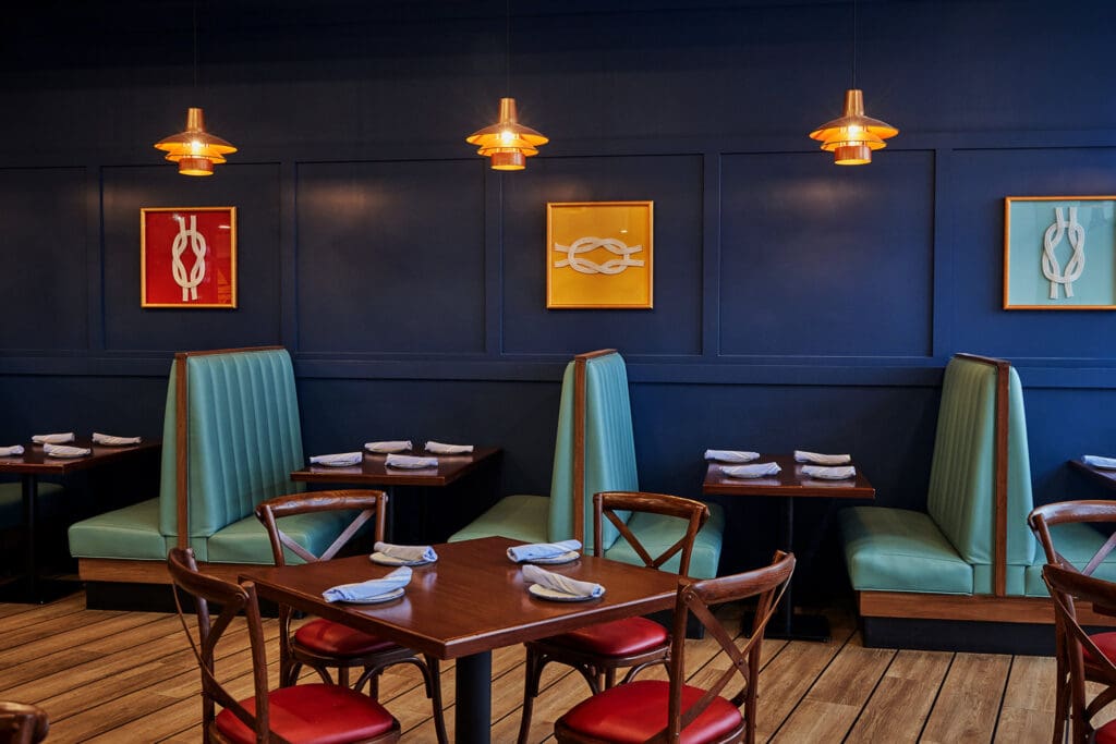 A cozy restaurant interior with blue walls, wooden flooring, and green vinyl booth seating. Wooden tables have red cushioned chairs and are set with neatly folded napkins. Three pendant lights hang from the ceiling, illuminating colorful wall art.