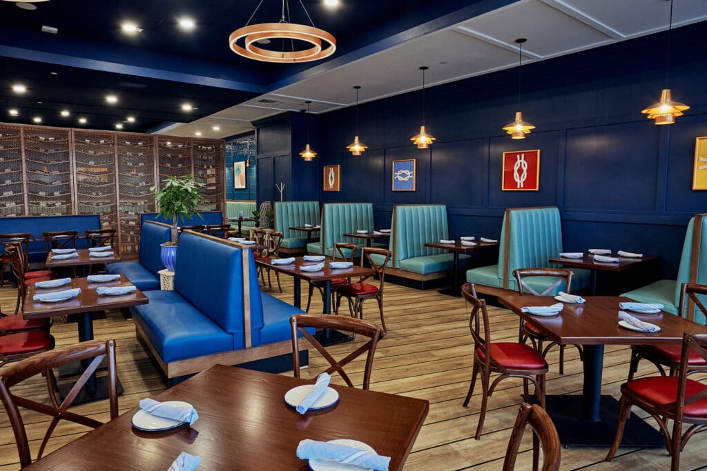 A modern restaurant with dark blue walls and wooden floors. The space features both booth and table seating with teal cushions and dark wooden chairs with red seats. Pendant lights hang from the ceiling, and framed art decorates the walls. Tables are set with white napkins.