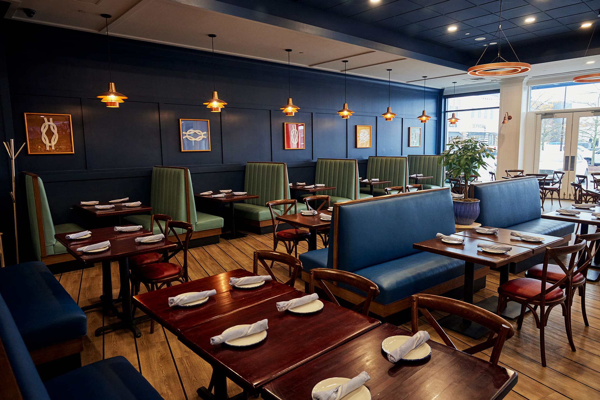 A modern restaurant interior with wooden floors, green and blue cushioned booth seating, and dark wood tables. Each table is set with white plates and napkins. Pendant lights hang from the ceiling, and framed art decorates the dark blue walls.