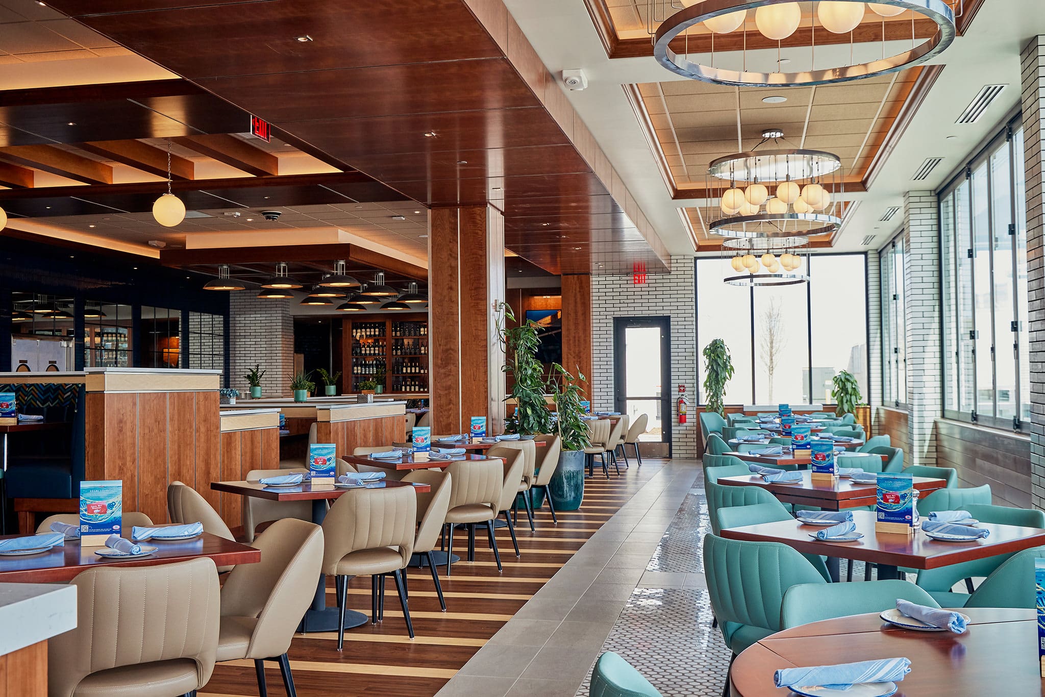 A modern, well-lit restaurant interior with wooden accents. There are multiple tables set for dining with blue napkins and menus. Large windows along one side let in natural light. Circular light fixtures hang from the ceiling. Green plants are placed around the space.