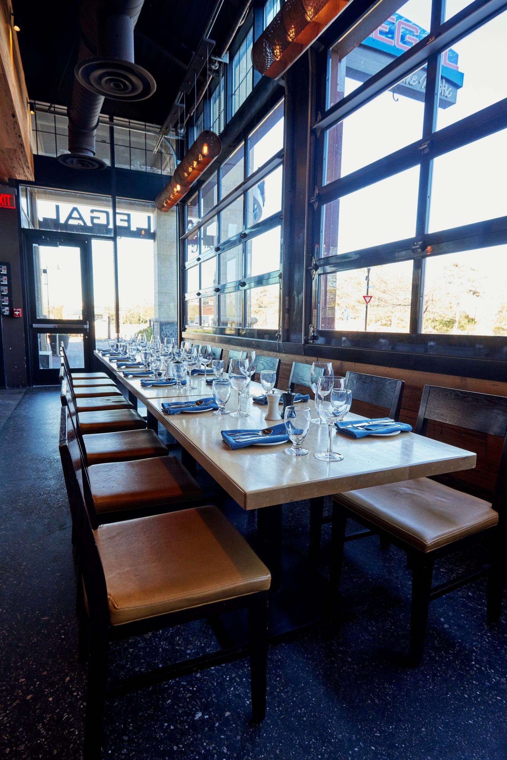 A modern restaurant interior featuring a long, rectangular table set with blue napkins, wine glasses, and water glasses. Wooden chairs with cushioned seats line either side of the table. Large windows and an industrial ceiling create an airy, well-lit atmosphere.