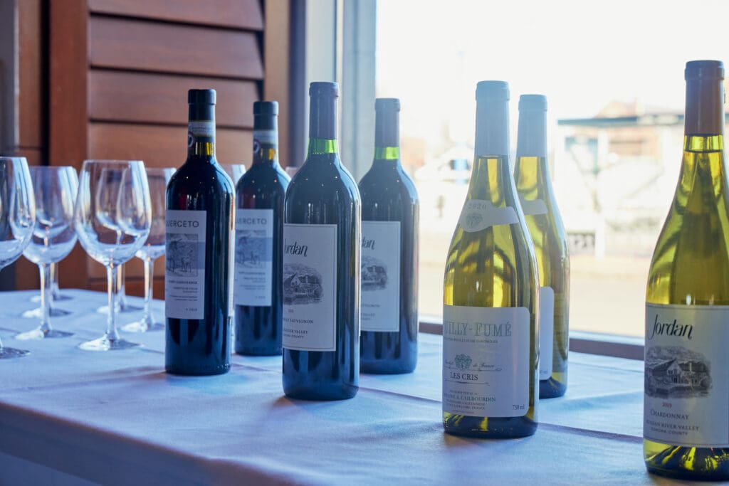 A white tablecloth holds several bottles of red and white wine alongside four empty wine glasses. The scene is set against a sunny window, revealing a view of the outside. Labels on the bottles show various wine brands and types.