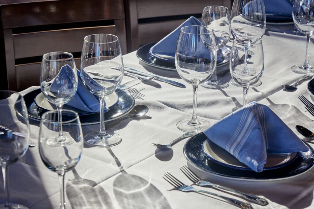 A dining table is elegantly set with blue napkins folded into triangles on top of white plates. Wine glasses and water glasses are placed alongside silverware on a white tablecloth, ready for a meal. The setting is sunlit.