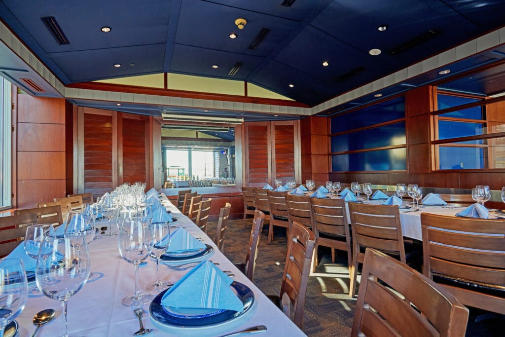 Brightly lit, elegant restaurant dining area with a long table set for guests. The table is adorned with white tablecloths, blue napkins, and glassware. Wooden chairs and paneling adorn the space, and a view of a bar area is visible in the background.