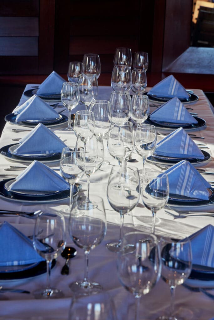 A dining table is elegantly set with blue napkins folded into triangles, placed on white plates. Surrounding each setting are multiple wine glasses, with sunlight casting shadows on the white tablecloth, creating a formal and sophisticated ambiance.