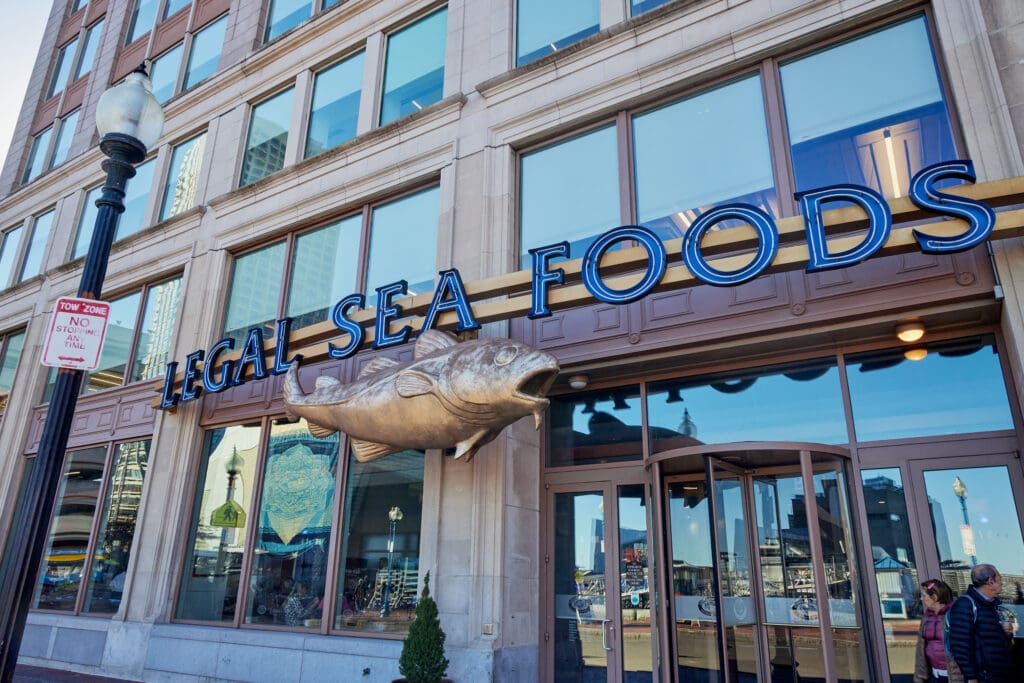 Storefront of Legal Sea Foods restaurant featuring a large fish sculpture above the entrance. The building has large windows and a brick facade. An adjacent street lamp has a 