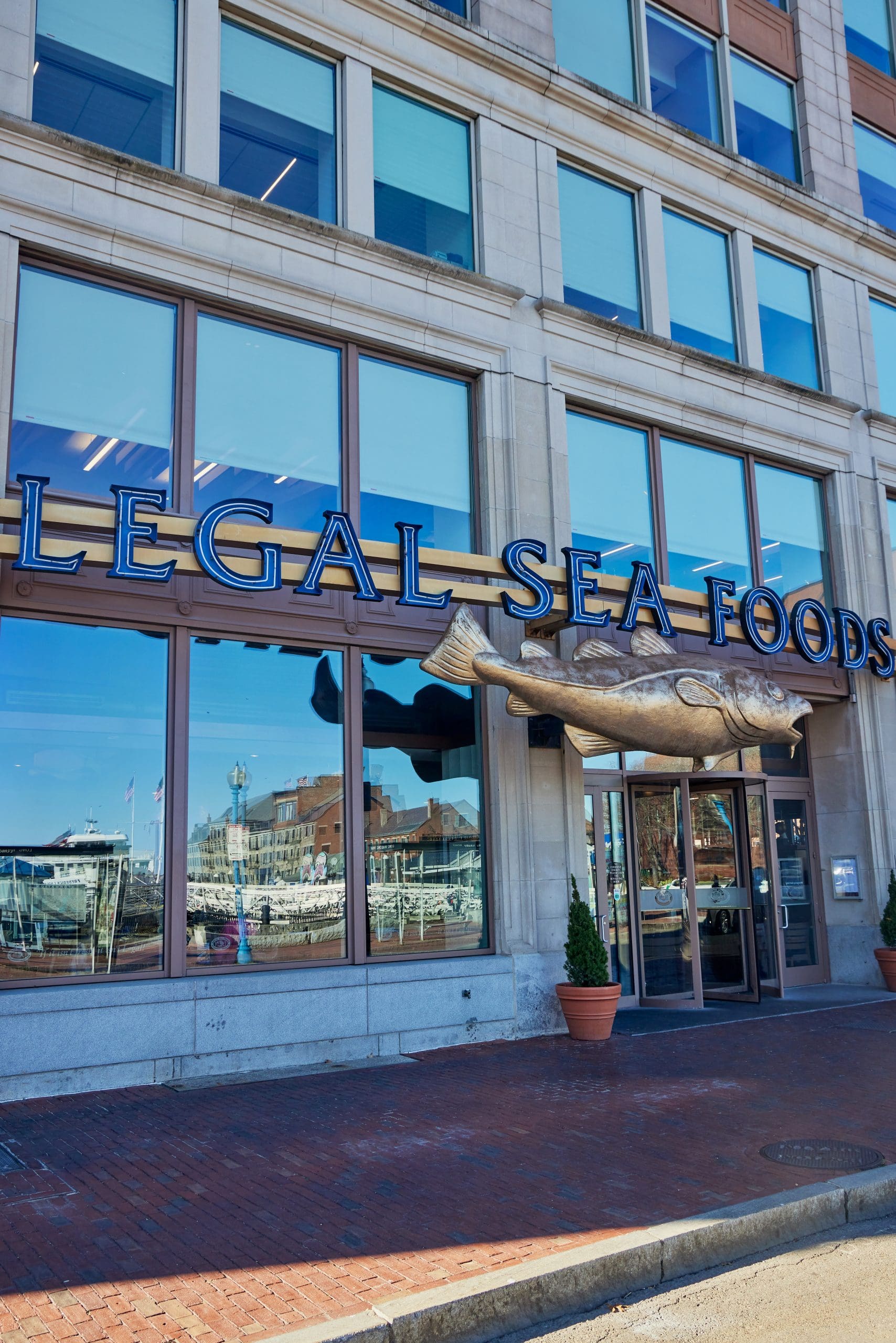 The image shows the entrance of a Legal Sea Foods restaurant. Large glass windows display reflections of nearby buildings, and the restaurant's name is prominently displayed above the door with a large fish sculpture attached to the sign.