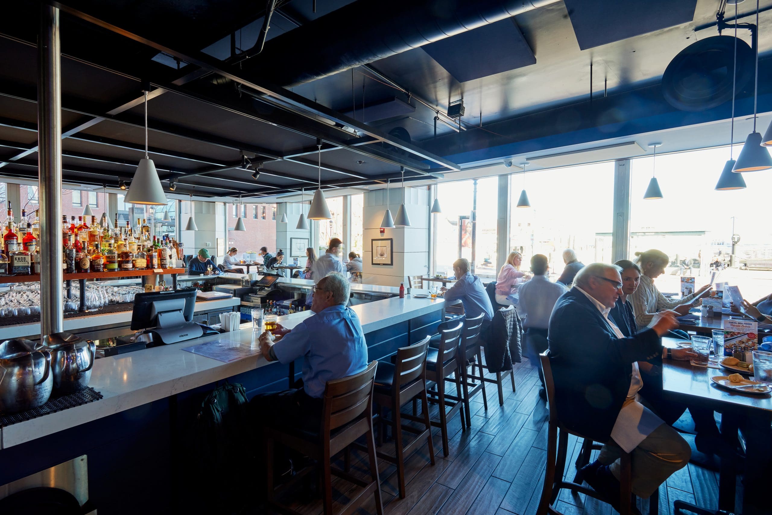 A dimly lit bar and seating area with a long, white counter and several bar stools. Patrons, mostly in casual or business attire, sit at the bar and tables near large windows that let in natural light. Bottles and glasses are arranged behind the bar.