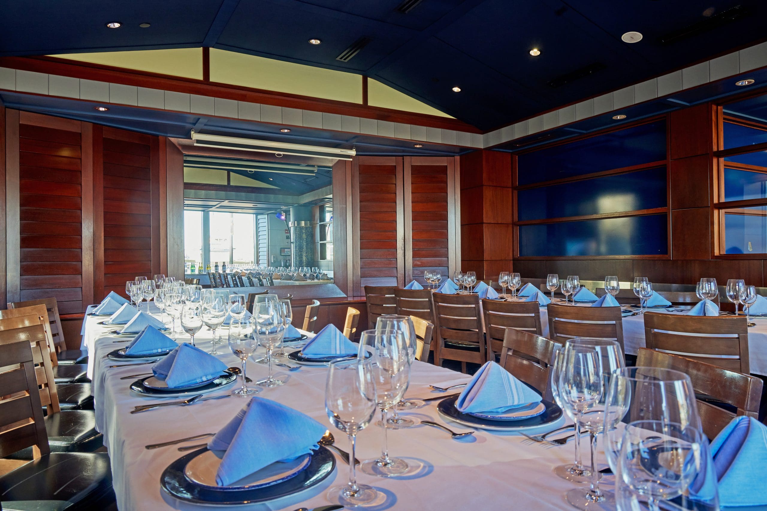 A dining area is set up for a formal event, with long tables draped in white tablecloths, set with blue napkins, wine glasses, and silverware. Wooden chairs surround the tables, and the space features dark wood paneling and blue accents.