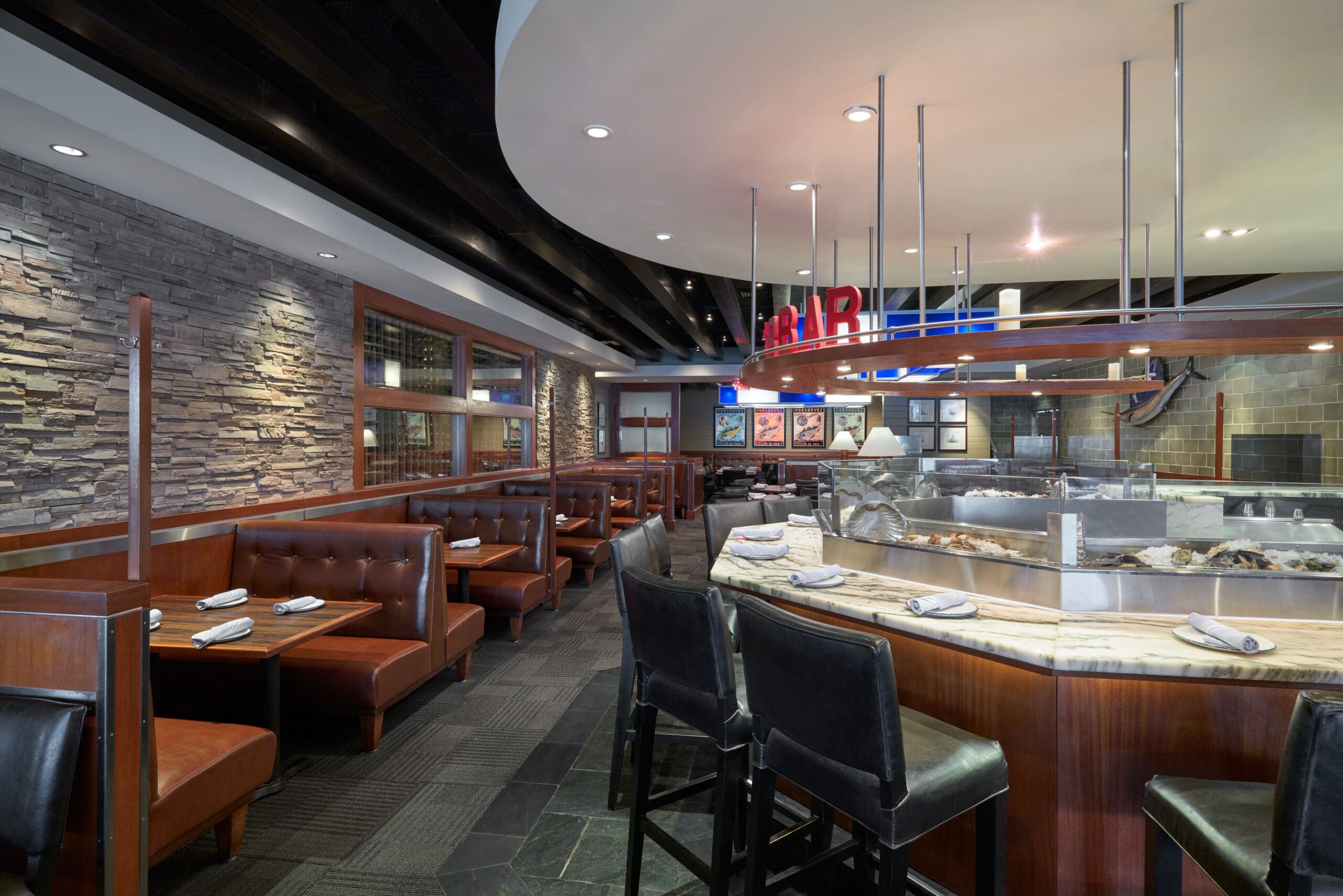 A modern restaurant interior featuring a mix of booth seating and a marble bar with high chairs. The booths are made of dark wood and upholstered in brown leather. A stone accent wall enhances the ambiance. Rolls of napkins are placed on the tables and bar.