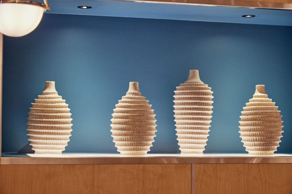 Four ribbed ceramic vases of varying heights are displayed on a shelf against a blue background, illuminated by soft lighting. The vases have a natural, earthy texture and pattern.