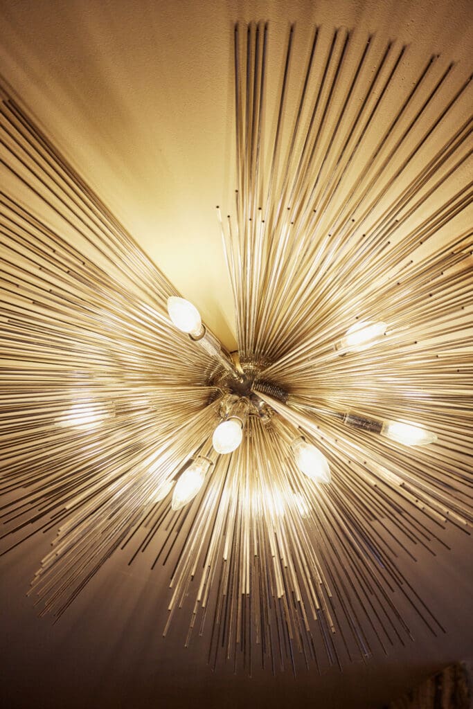 Close-up view of a modern, starburst-style ceiling light fixture with multiple bulbs and metallic rods radiating in all directions, creating a dynamic and visually striking design against the ceiling.