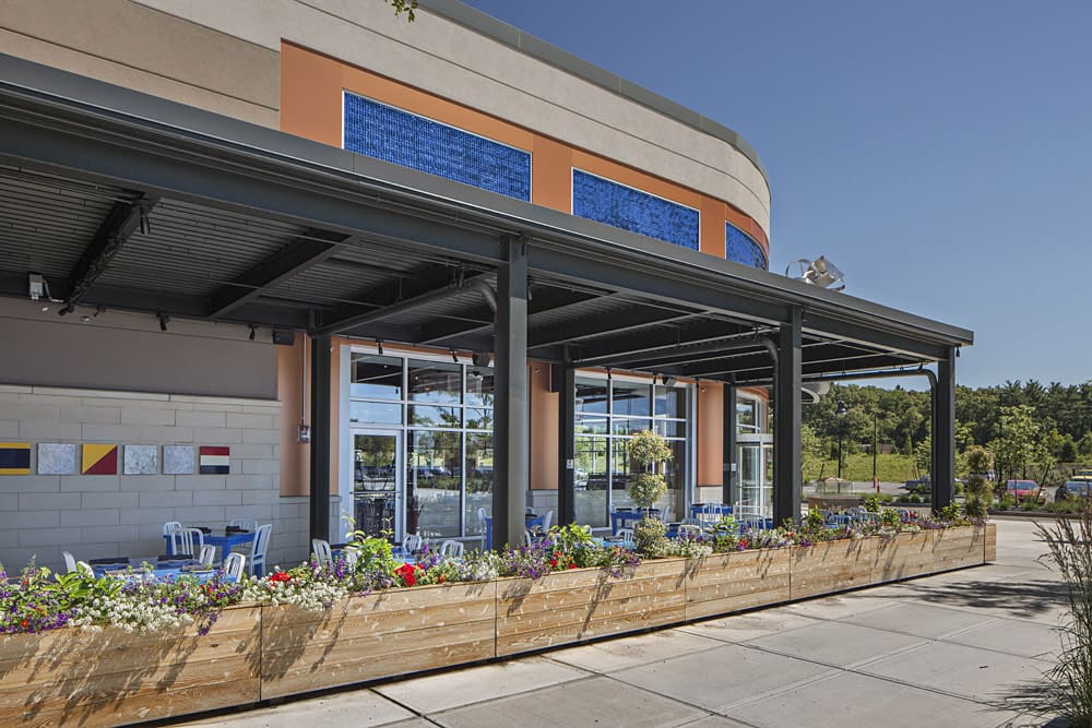 Outdoor seating area of a modern café or restaurant with a covered pergola and potted flowers in wooden planters. The building features a mix of large glass windows and blue accent tiles. Trees and greenery are visible in the background under a clear sky.