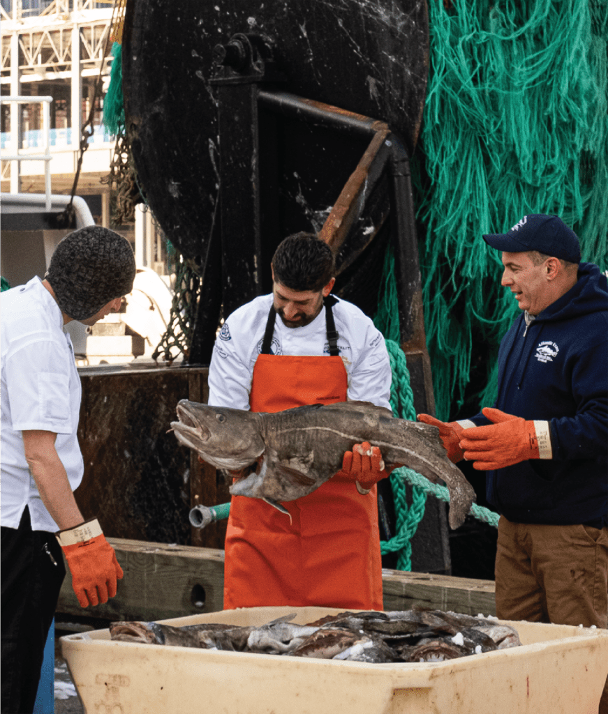 Three fishermen are standing, two in white outfits and one in a blue jacket, holding a large fish. One man is presenting the fish to the others. They are on a dock with a large net hanging in the background and a container filled with more fish in the foreground.