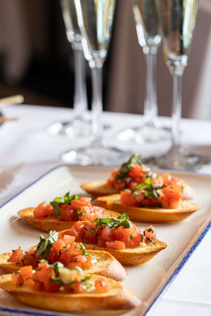 A white rectangular plate holds six pieces of bruschetta topped with diced tomatoes and fresh herbs. The plate sits on a white tablecloth, accompanied by three tall glasses of champagne in the background. The setting suggests a fine dining environment.