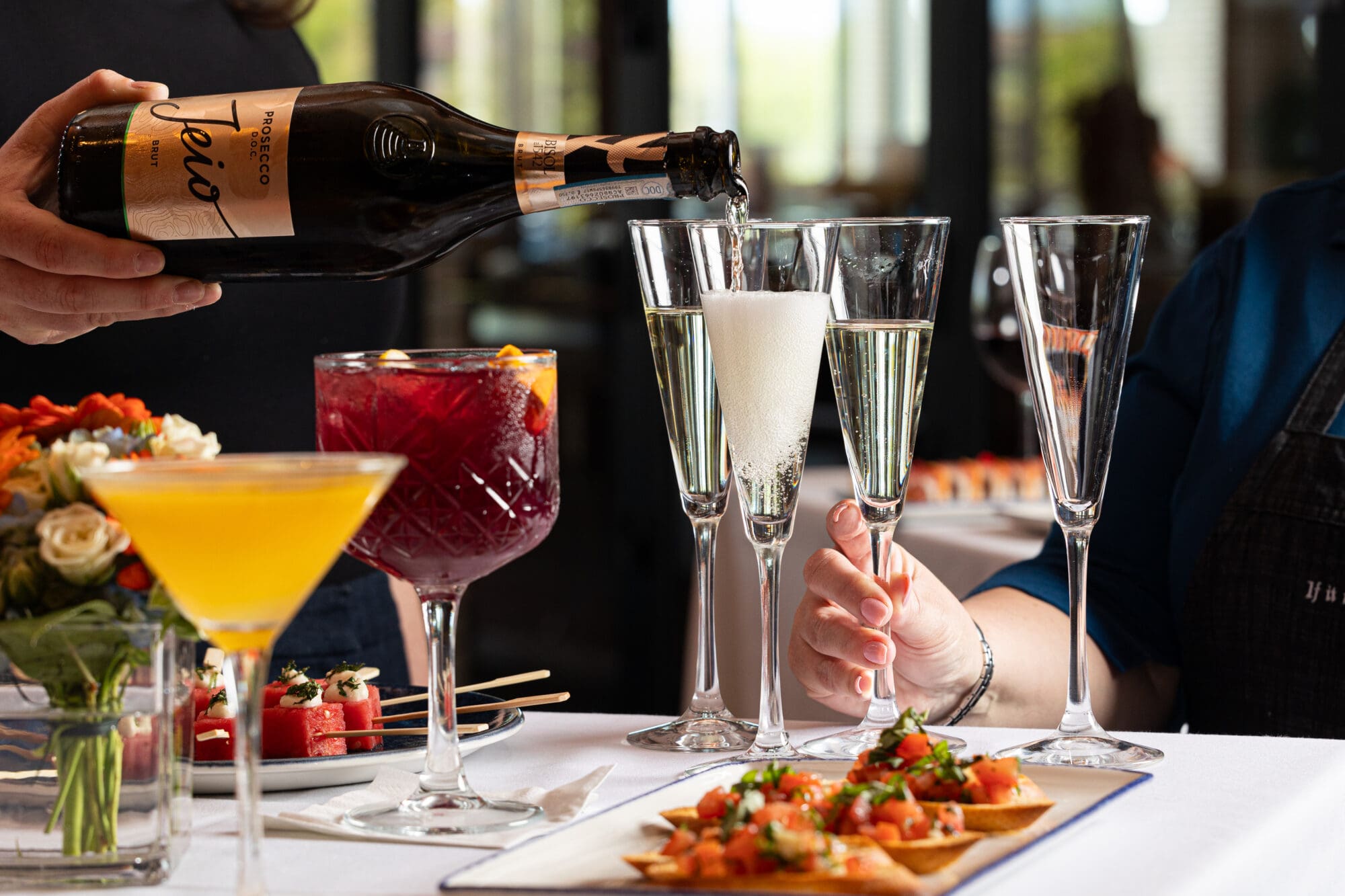 A person pours prosecco into a champagne flute at a table adorned with a variety of colorful cocktails and appetizers. The setting appears to be a sophisticated indoor gathering with a focus on fine dining.