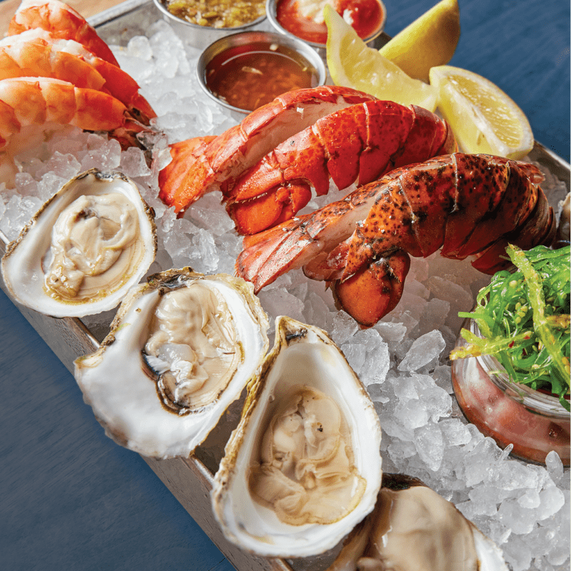 A platter of seafood featuring oysters on the half shell, lobster tails, and large shrimp arranged on a bed of ice. Sauces, lemon wedges, and a side of greens accompany the seafood, all presented on a blue surface.