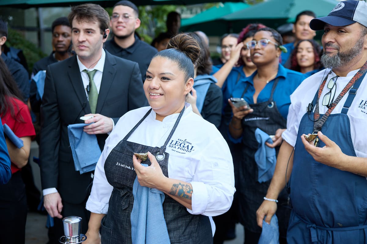 A group of people gathered outdoors, featuring a woman in a chef's uniform holding a small dish and a man in a similar uniform holding a towel. Others in the crowd, wearing business or casual attire, appear to be watching something and holding blue napkins.