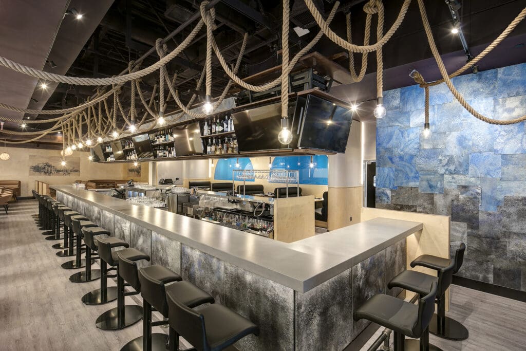 A modern bar with a sleek metallic counter, black barstools, and multiple TVs mounted above. Ropes and hanging light bulbs adorn the ceiling. The walls feature abstract blue and silver designs, adding to the contemporary ambiance. The space is empty of people.