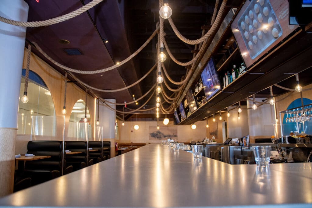 A modern bar with a maritime theme features hanging rope lights and wooden accents. The bar counter is lined with empty glasses and overlooks a spacious seating area with booth seating. The ceiling is adorned with additional rope decor, creating a cozy ambiance.