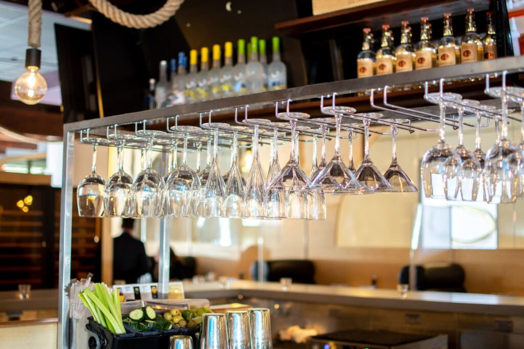 A bar area with various glasses hanging upside down from a rack. Bottles of liquor are displayed on shelves above the glasses. Bar tools and ingredients, including limes, are placed on the counter below. The background shows a seating area with tables and chairs.