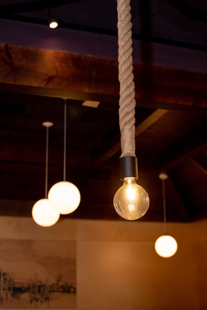 A hanging light bulb with a rope-wrapped cord is illuminated in the foreground. In the background, three spherical lights are softly glowing, suspended from the dark wooden ceiling, creating a warm and cozy ambiance.