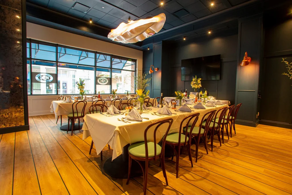 A sophisticated dining room with tables covered in white tablecloths and set for a meal, complete with wine glasses, folded napkins, and floral centerpieces. The room has wooden floors, dark wall paneling, a large window, and modern overhead lighting.