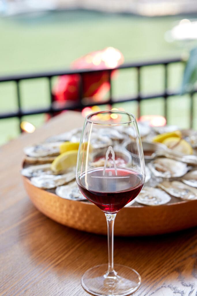 A glass of red wine sits on a wooden table, with a plate of oysters and lemon wedges in the background. The scene is set outdoors, with a blurred view of water, railing, and some lights, suggesting a peaceful waterfront dining experience.