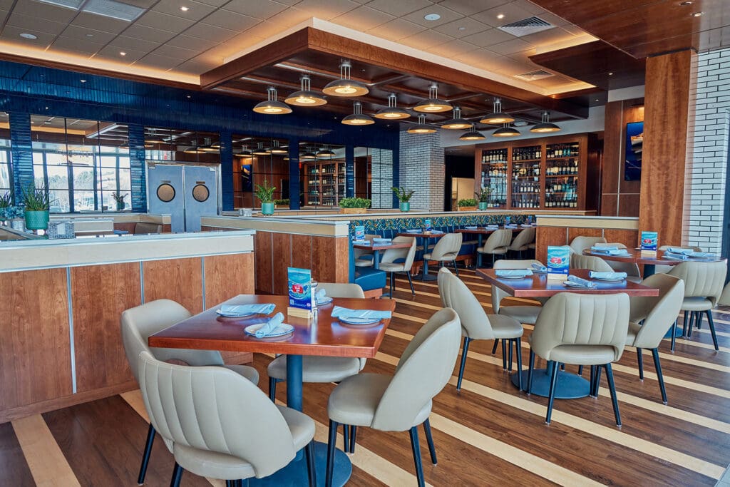 A modern restaurant interior featuring wooden tables and cushioned beige chairs. The space is well-lit with ceiling lights, and there are plants adorning some surfaces. In the background, shelves filled with wine bottles can be seen, and large windows let in natural light.