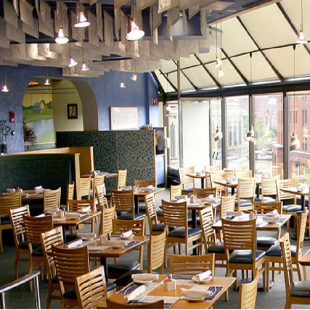 A modern, spacious restaurant with large windows offering city views, filled with wooden tables and chairs. Tables are set with white napkins and silverware. Overhead, unique ceiling decor features hanging panels. The setting is bright with ample natural light.