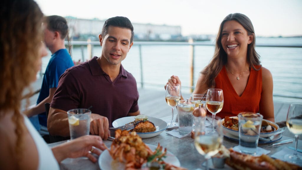 A group of friends enjoys a meal at an outdoor waterfront restaurant. A man in a maroon shirt and a woman in a red top are smiling and talking, while the table is set with elegant dishes and glasses of white wine. The background features a scenic water view.