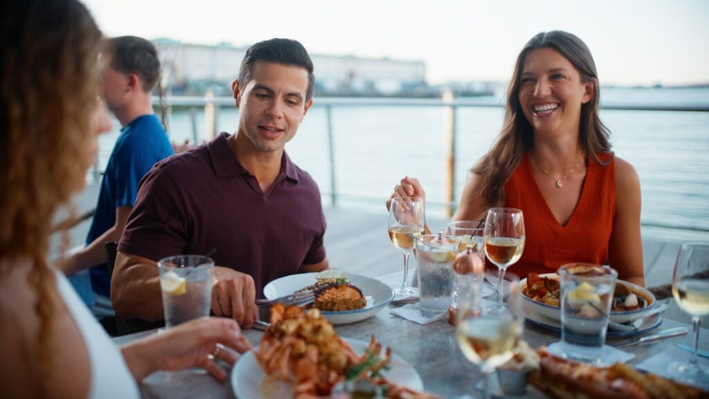 A group of three people sits at an outdoor restaurant table overlooking the water. They are enjoying a meal with drinks, smiling and engaging in conversation. Various dishes and glasses of wine are on the table, and the background shows a scenic waterfront.